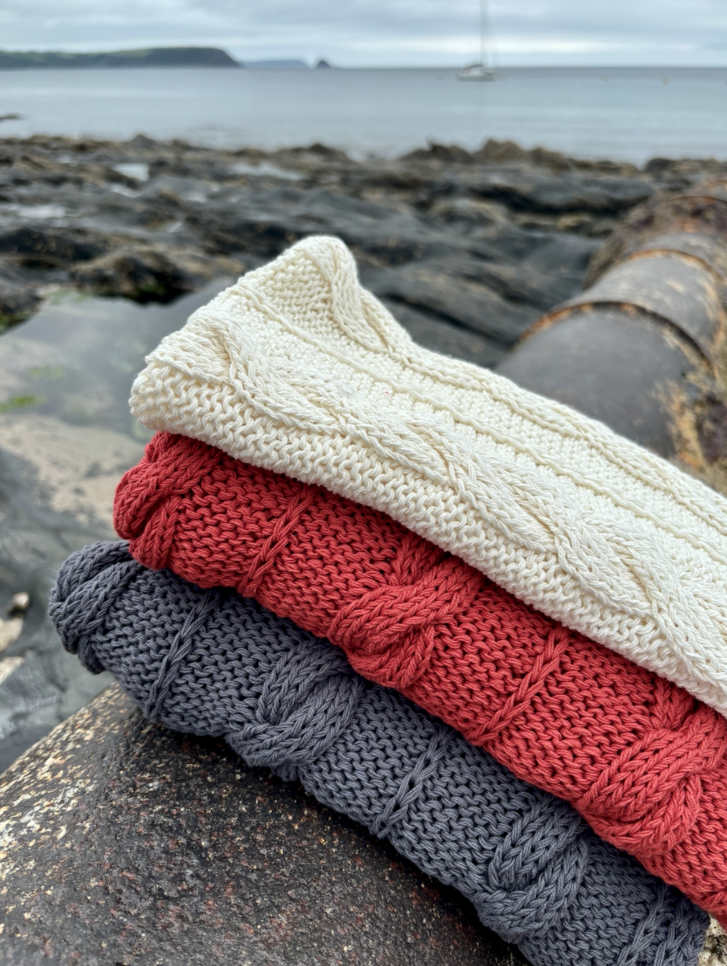 A stack of hand knitted baby blankets in beige, gray, and red, placed near a rocky shoreline with a boat in the background.