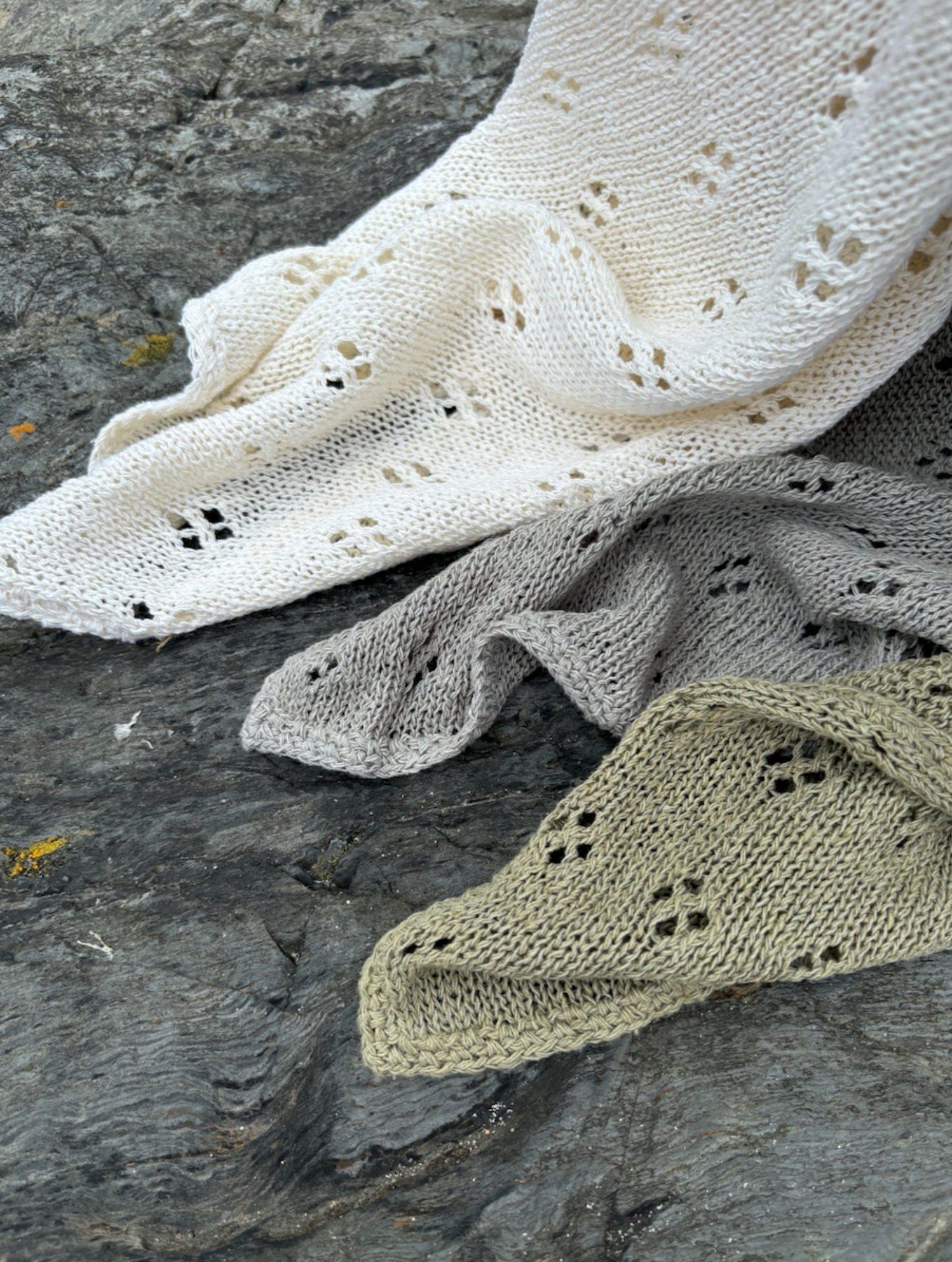 A selection of hand knitted baby blankets in creme, storm blue grey, and coriander green, placed on a rocky shoreline