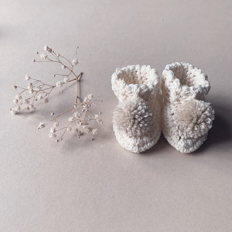 Image shows a pair of organic cotton cream baby booties with oatmeal pom pom